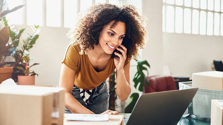 Junge Frau telefoniert mit dem Handy, während Sie einen Laptop bedient zwischen Umzugskartons