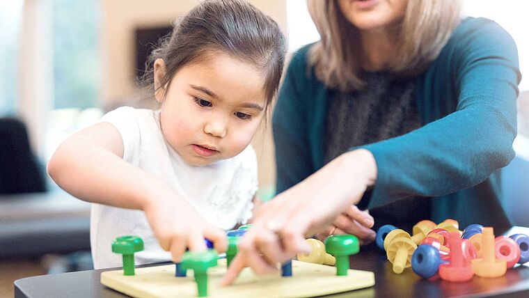 Ergotherapie-Blankoverordnung: Ein Mädchen spielt mit ihrer Ergotherapeutin ein Spiel mit bunten Knöpfen.