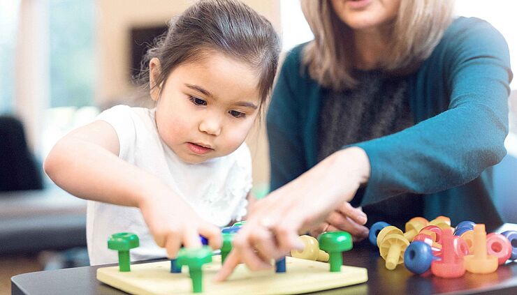Ergotherapie-Blankoverordnung: Ein Mädchen spielt mit ihrer Ergotherapeutin ein Spiel mit bunten Knöpfen.