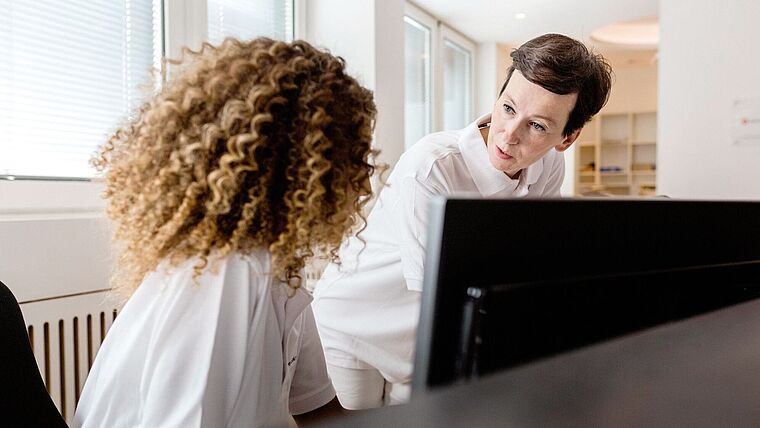 zwei weibliche Praxisverwaltungskräfte diskutieren hinter einem Tresen an einem Computerbildschirm