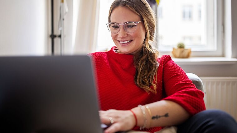 Junge Frau mit rotem Pullover sitzt zuhause am Laptop