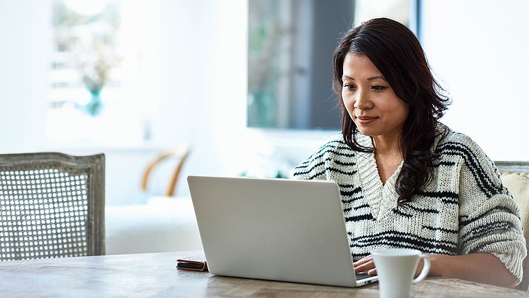 Frau mit schwarzen Haaren sitzt am Tisch mit einer Tasse Kaffee und ist an ihrem Laptop.