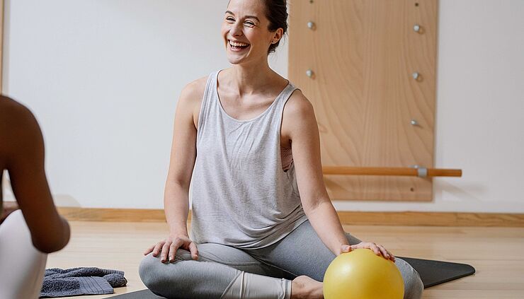 Fröhliche Frau in Sportkleidung sitzt auf einer Yogamatte eines Trainingsbereichs in der Physiotherapie